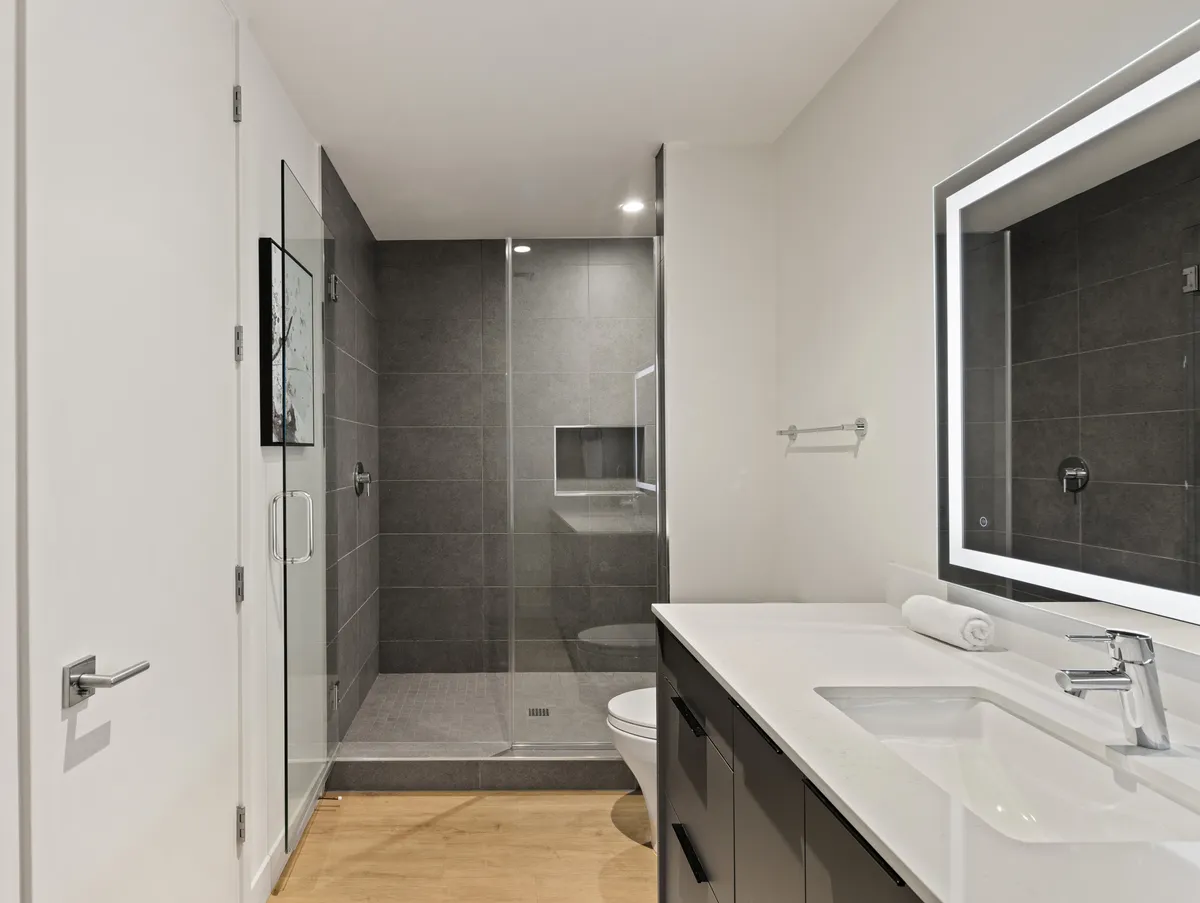 A modern ensuite bathroom with a striking glass shower enclosure, elegant vanity, and a large backlit mirror. The overall design is contemporary, serene, and spa-inspired, offering a glimpse into the premium bathroom experience available in the bedrooms.