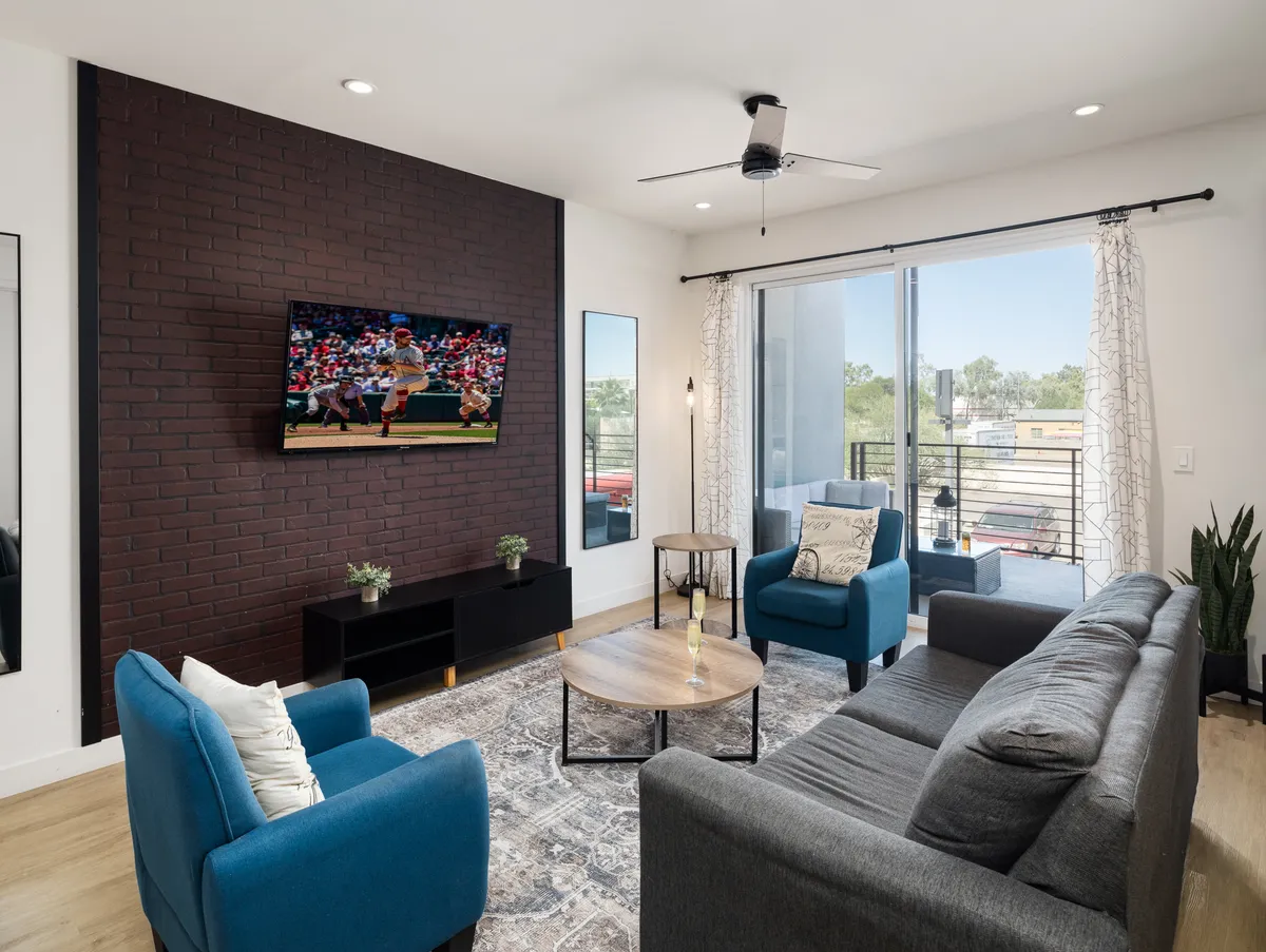 The image showcases a stylish, modern living room interior featuring comfortable blue accent chairs arranged around a sleek coffee table, facing a large TV displaying a sports game. The atmosphere is inviting, perfect for guests looking to relax, watch sports, or gather with friends in a cozy, open space.