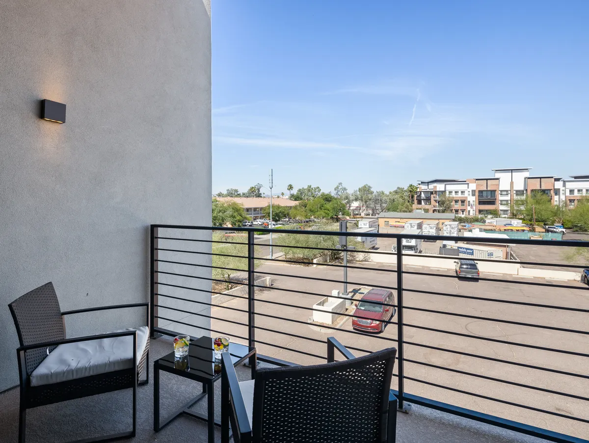 A serene, private balcony with comfortable seating and a view of the neighborhood. Glasses of drinks rest on the table, inviting relaxation and social moments outdoors. The scene captures both sunrise tranquility and the allure of sunset cocktails.