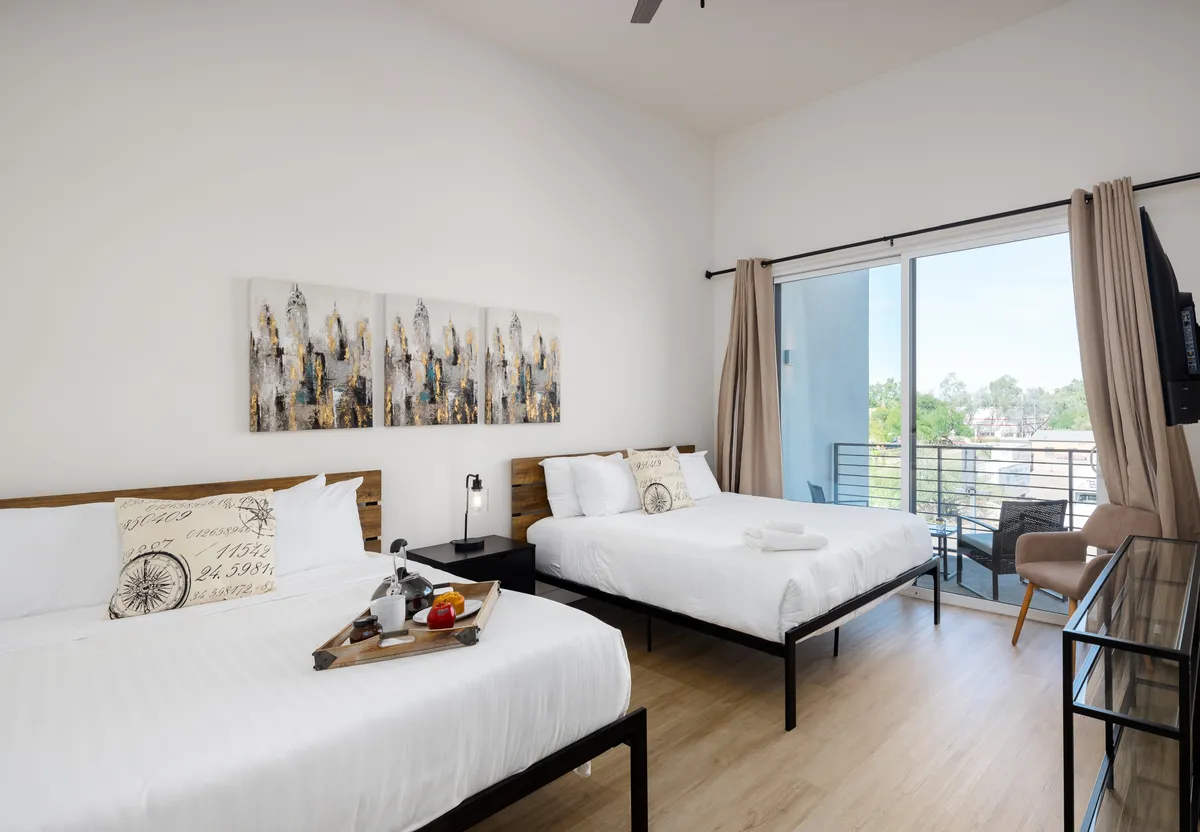 This image features a calming, contemporary bedroom with two Queen beds dressed in white linens, decorative wall art above the beds, and glass doors leading out to a private balcony. The room is spacious and sunlit, evoking a serene atmosphere ideal for relaxation. There is clear balcony access, hinting at the opportunity to step outside and greet the day.