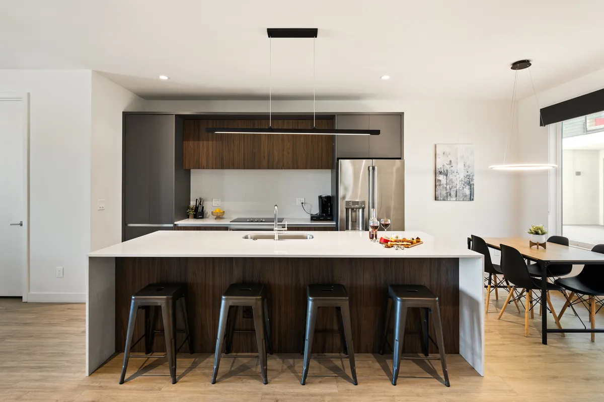 A sun-drenched open-concept kitchen, dining area, and living space. The kitchen features a large island with bar stools, glossy stainless steel appliances, and a contemporary dining table. The living area is visible nearby, creating a seamless flow for socializing and entertaining.