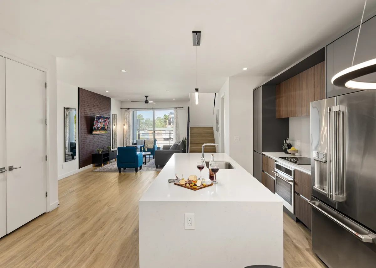 A modern kitchen island adorned with snacks and drinks, adjacent to a bright living area furnished with stylish blue chairs and flooded with natural light from large windows. The Smart TV is visible in the background, and both kitchen and living zones blend into one inviting communal space.