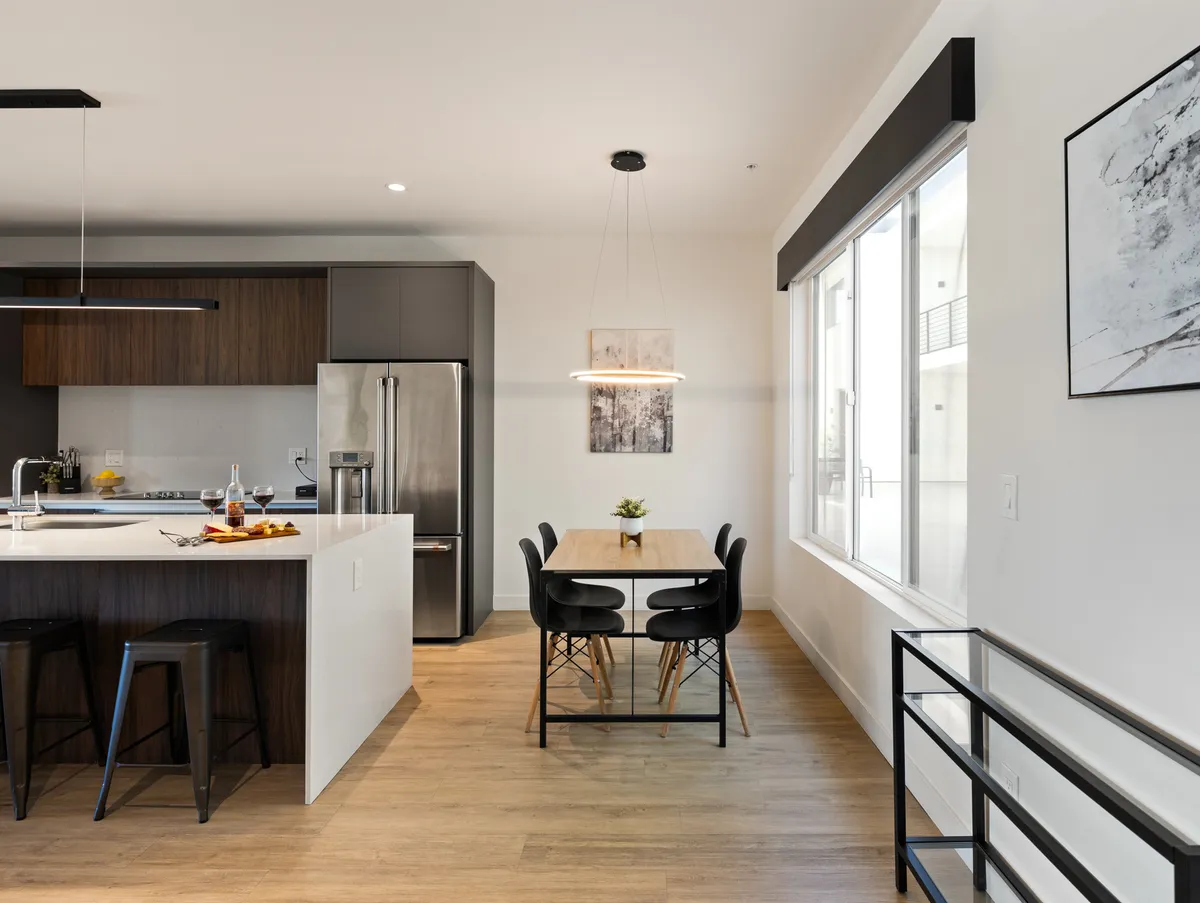 A welcoming kitchen and dining area with a spacious island countertop, adjacent dining table, and striking modern light fixtures overhead. The clean, contemporary style is complemented by soft sunlight streaming in, setting the scene for shared meals and conversations.