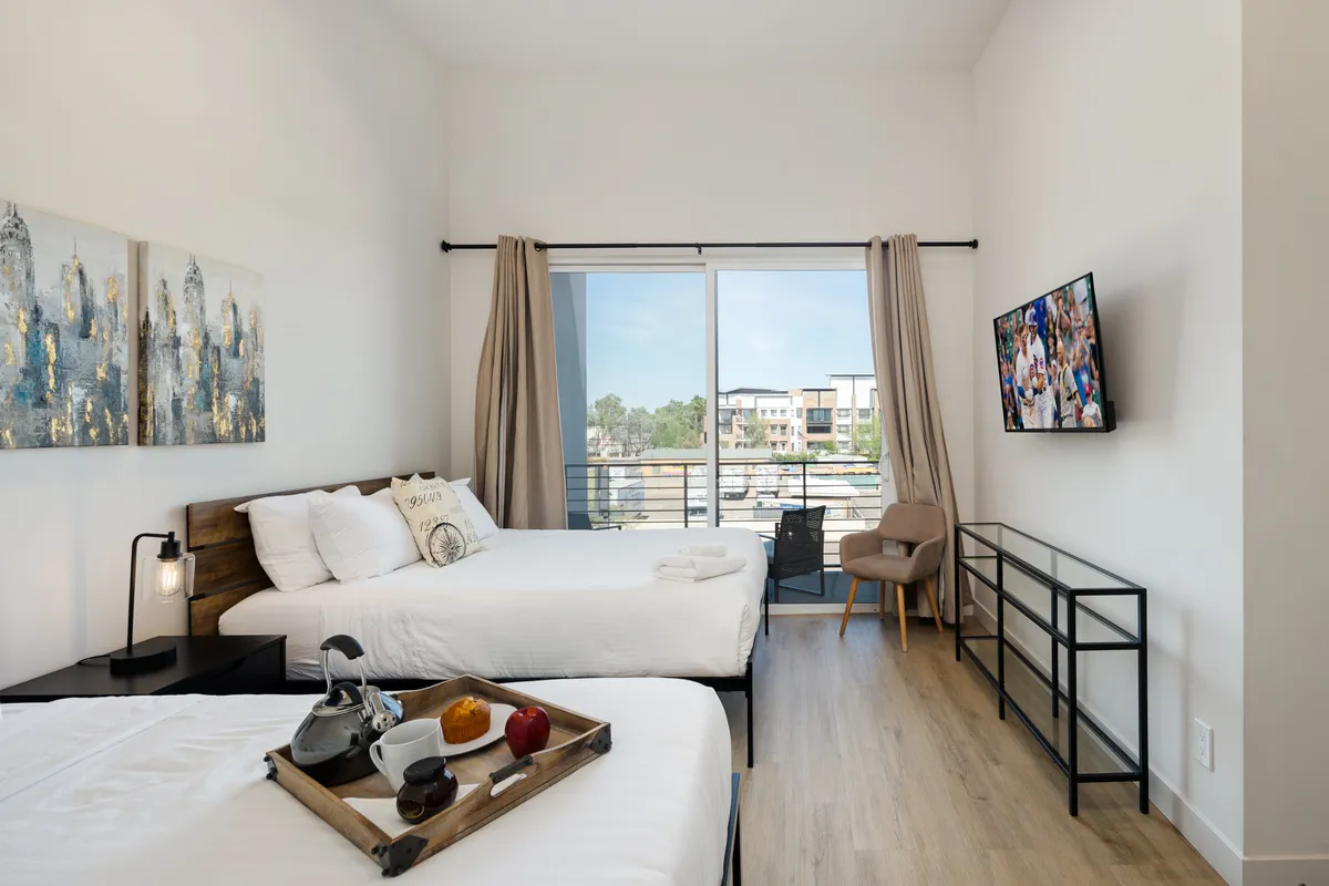 The bedroom showcases two Queen beds made up with crisp white linens, stylish wall art, a tray with breakfast items placed on one of the beds, and a large window offering natural light. The inviting setup suggests a space ideal for unwinding, socializing, and enjoying quiet mornings.